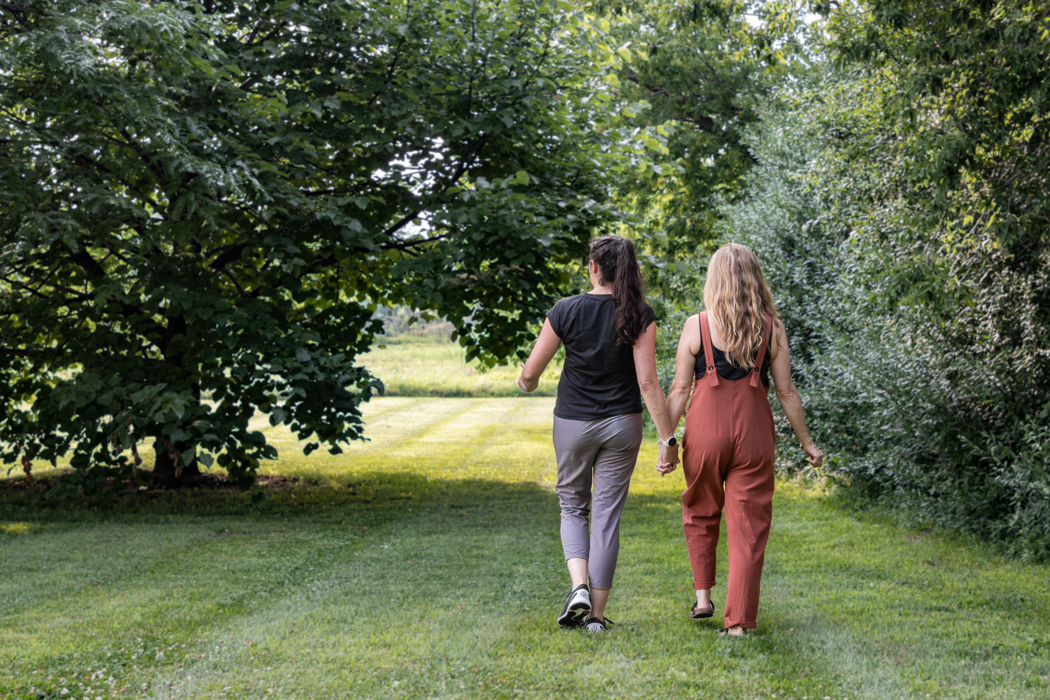 Two women walking hand in hand through a lush green park, surrounded by trees and grass, symbolizing support and connection.